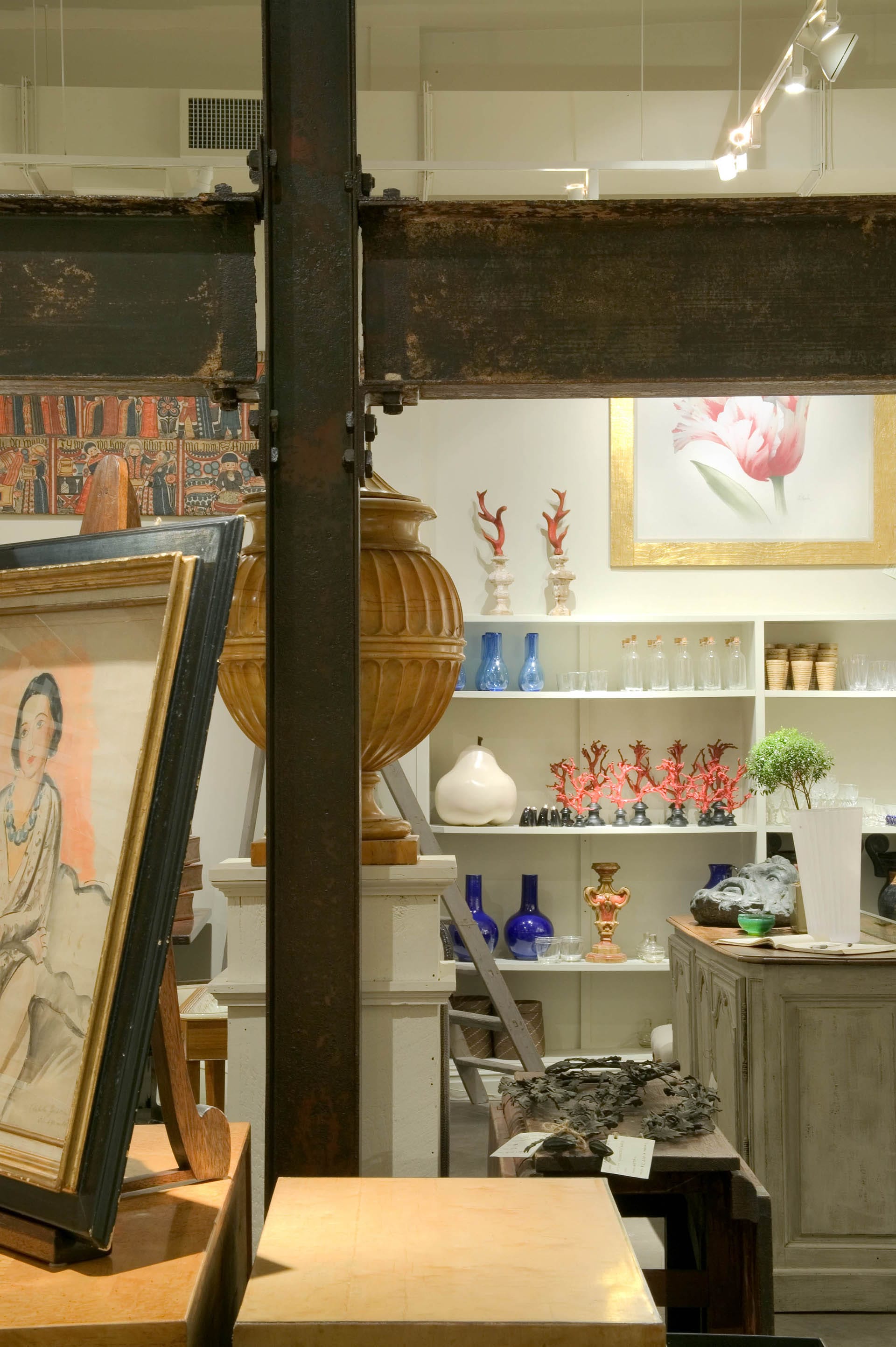 At the foreground, a wood table holds merchandise in an antique store. Beyond that sit decorative and supportive aged metal beams. In the background, a set of shelves holds antique glassware and decorative objects.