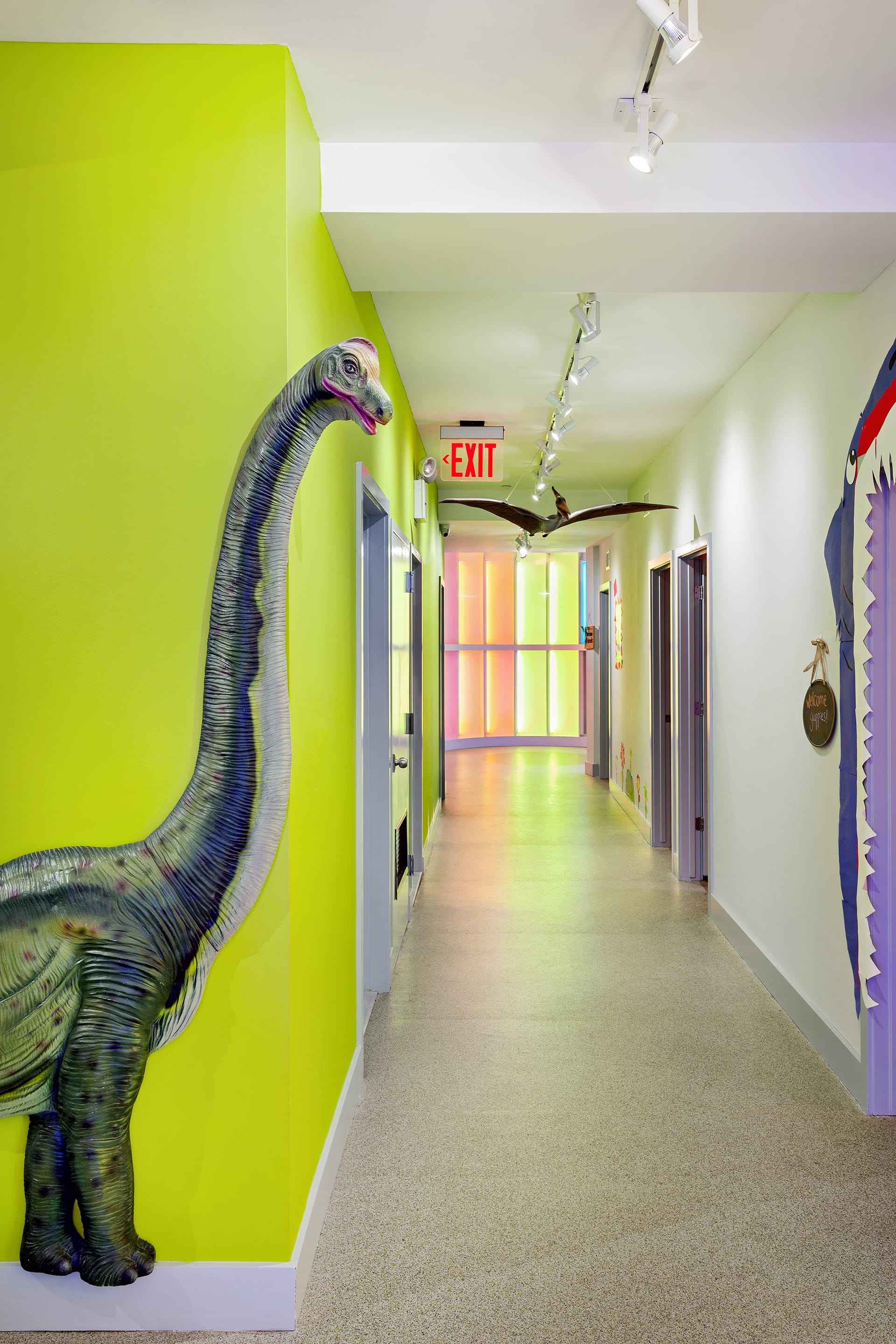 Hallway with bright green walls, a large dinosaur model, and a neon-lit rotunda at the end of the hall.