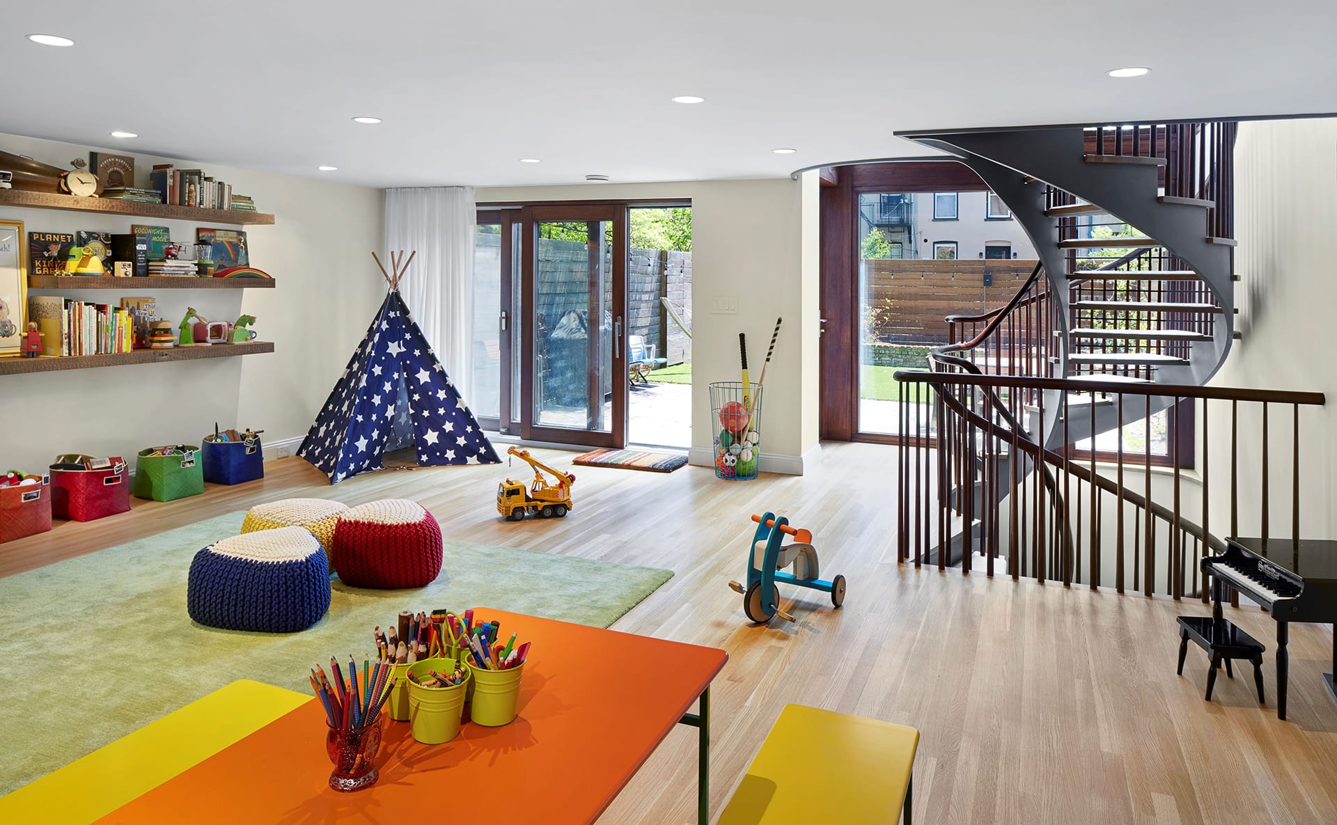 Playroom with a green carpet, orange and yellow picnic table, blue star-patterned teepee, open-riser and open stringer sculptural staircase, and dark wood accents. A child-size grand piano sits at the right corner of the frame.