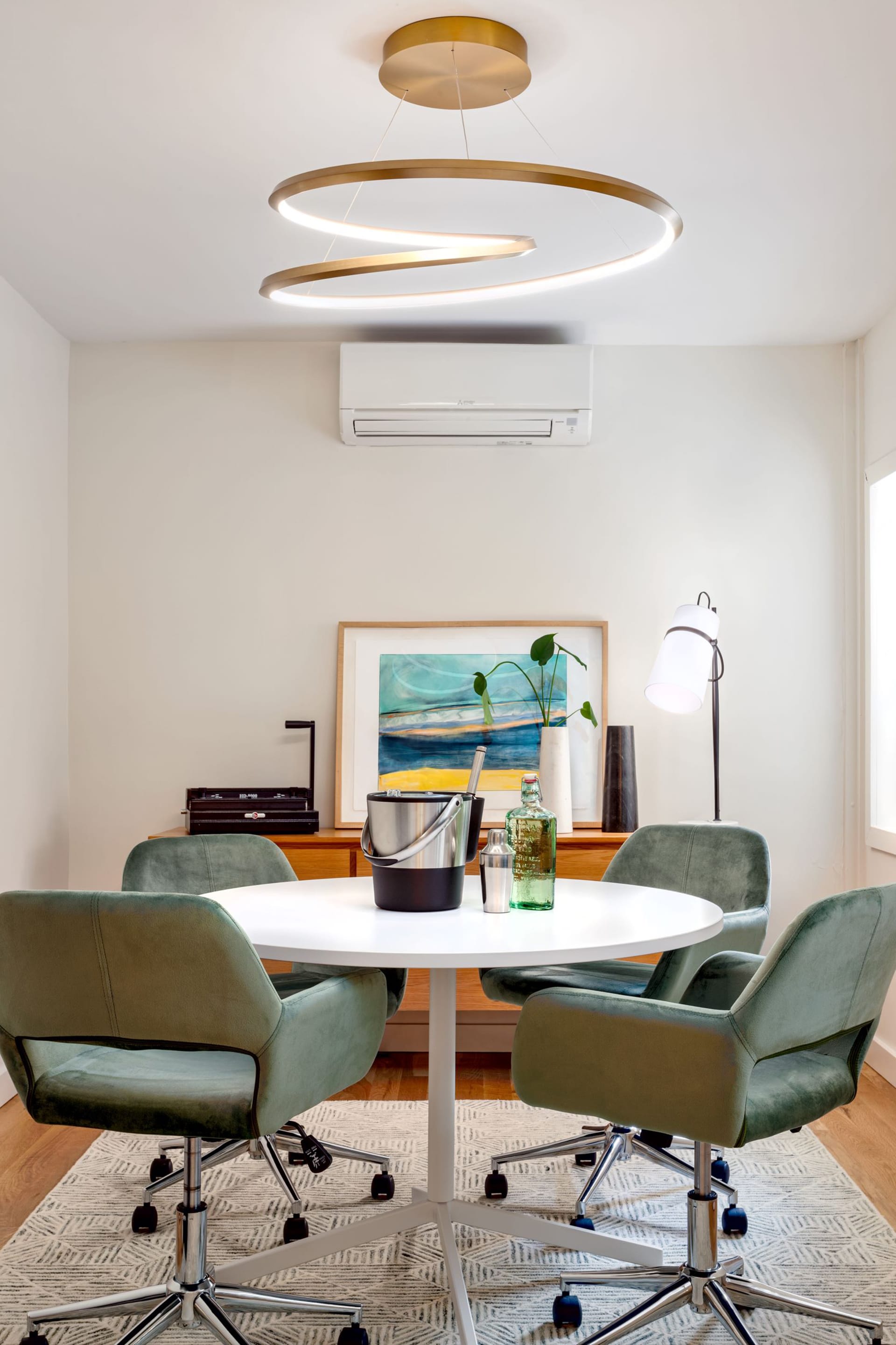 Office space with an abstract gold chandelier hanging above a round conference table with four green velvet chairs. Bar accoutrements sit atop the conference table, and an area rug sits below it.