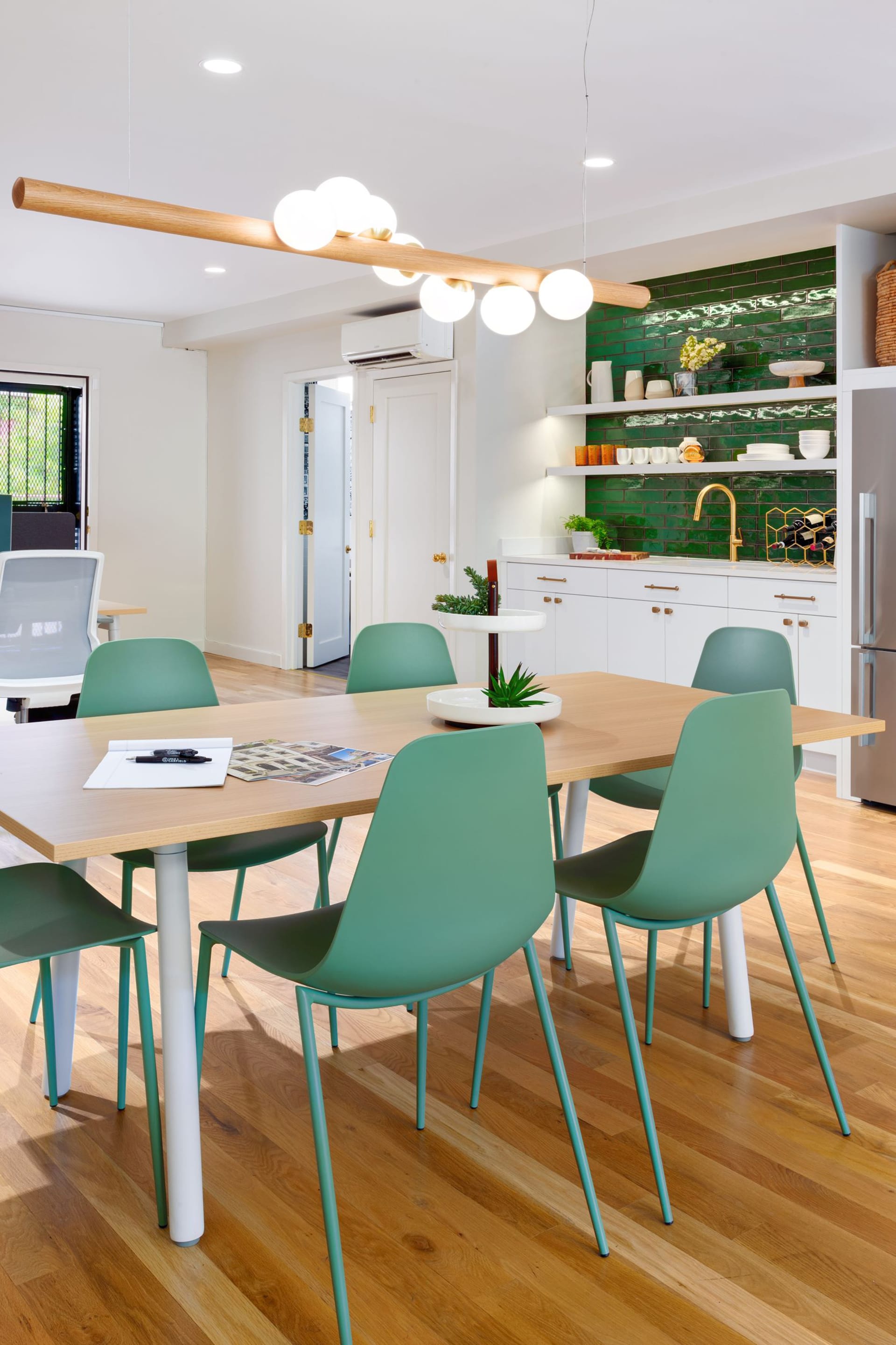 Dining area in a real estate office with wood floors, wood dining table, green dining chairs, and statement chandelier above the table. In the background sits a kitchenette with dark green subway tile backsplash.