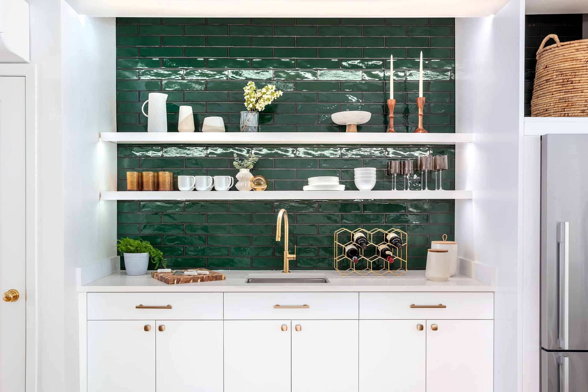 Kitchenette with gold fixtures, will millwork, green subway tiles, and various dishes and other objects.
