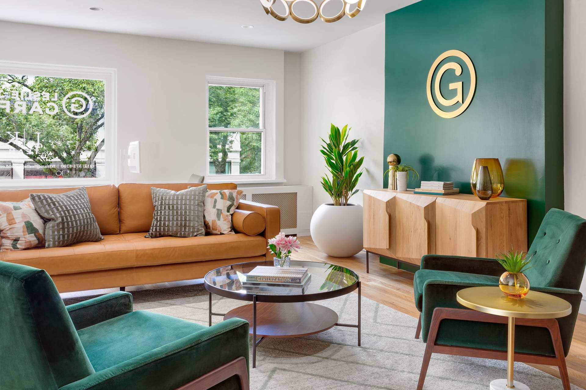 Lobby of a Brooklyn Heights real estate office with a green accent wall, wood credenza, two green velvet arm chairs, a glass coffee table, and a bright leather couch.