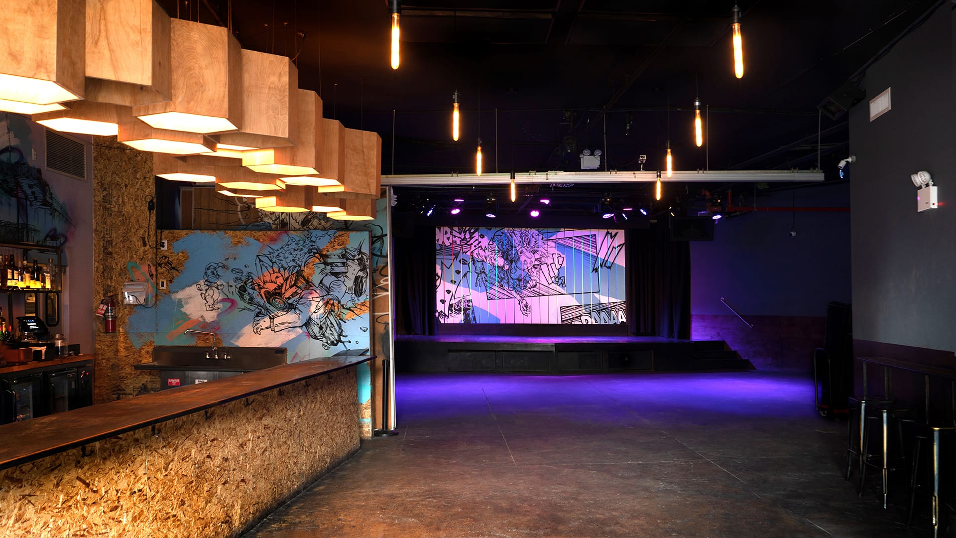 A large room containing a stage at the far end and a bar at the front left of the image. The bar is clad with cork, a mural lines the walls behind the bar, and a custom-made honeycomb light fixture hangs above it. Pendant lights in two straight lines lead back toward the stage, which has a custom-designed backdrop and lighting system.