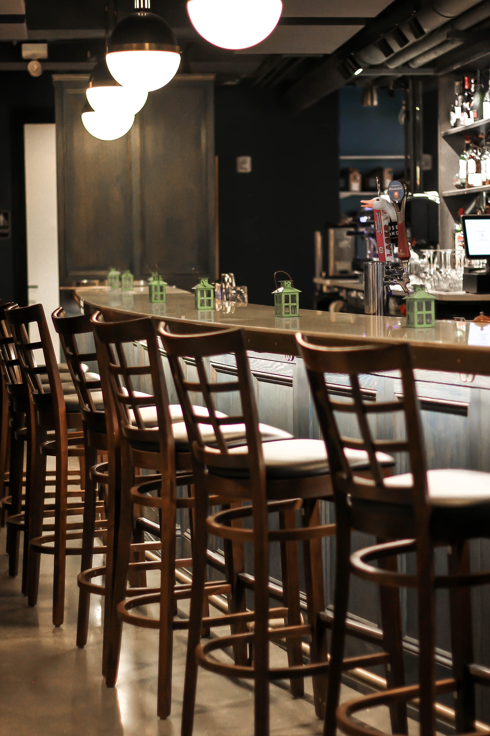 A row of barstools in front of a bar with green lanterns on top and alcove lighting underneath the bar.