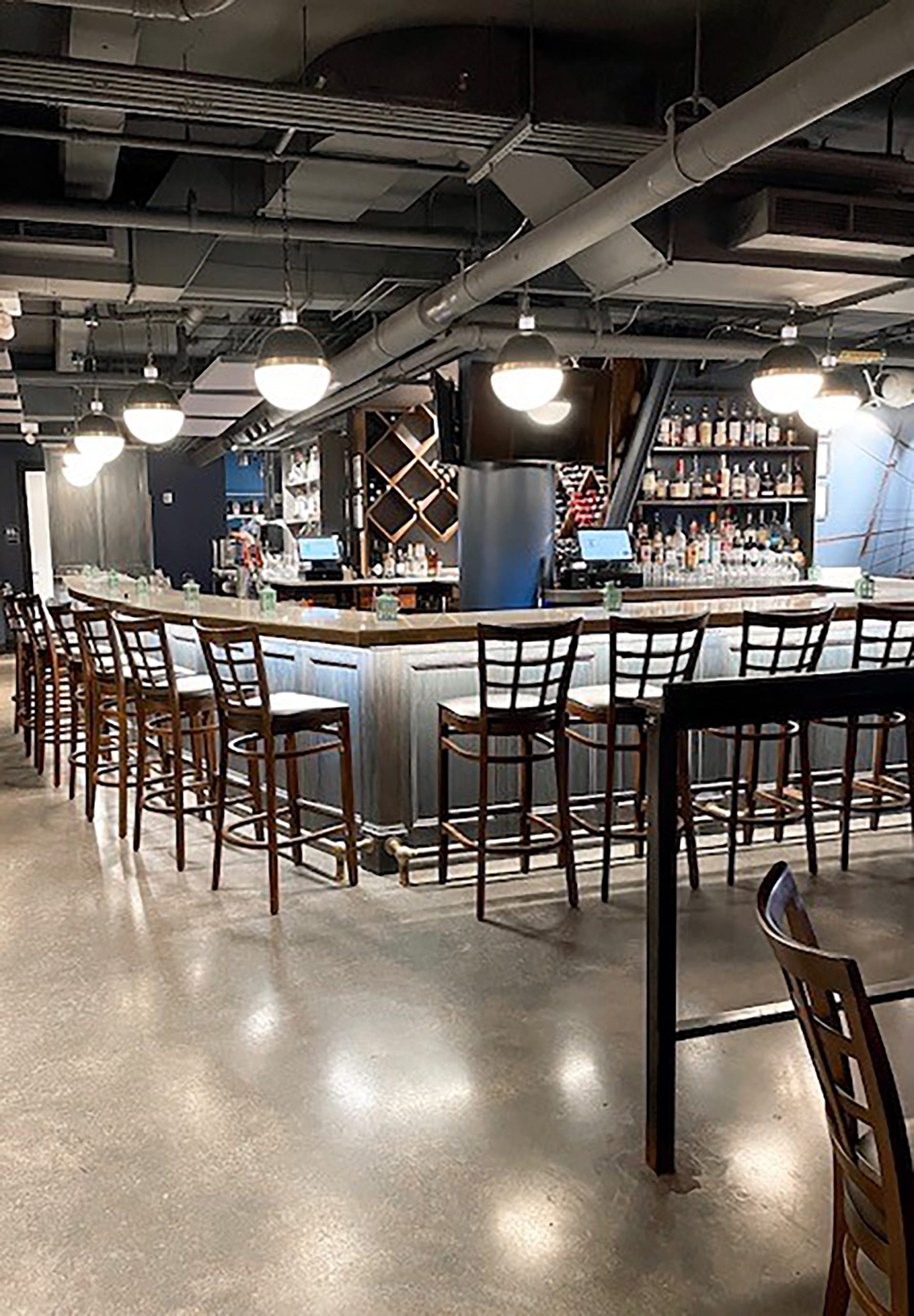 Bar with industrial roof, polished floors, under-bar lighting, and wooden barstools with backs.