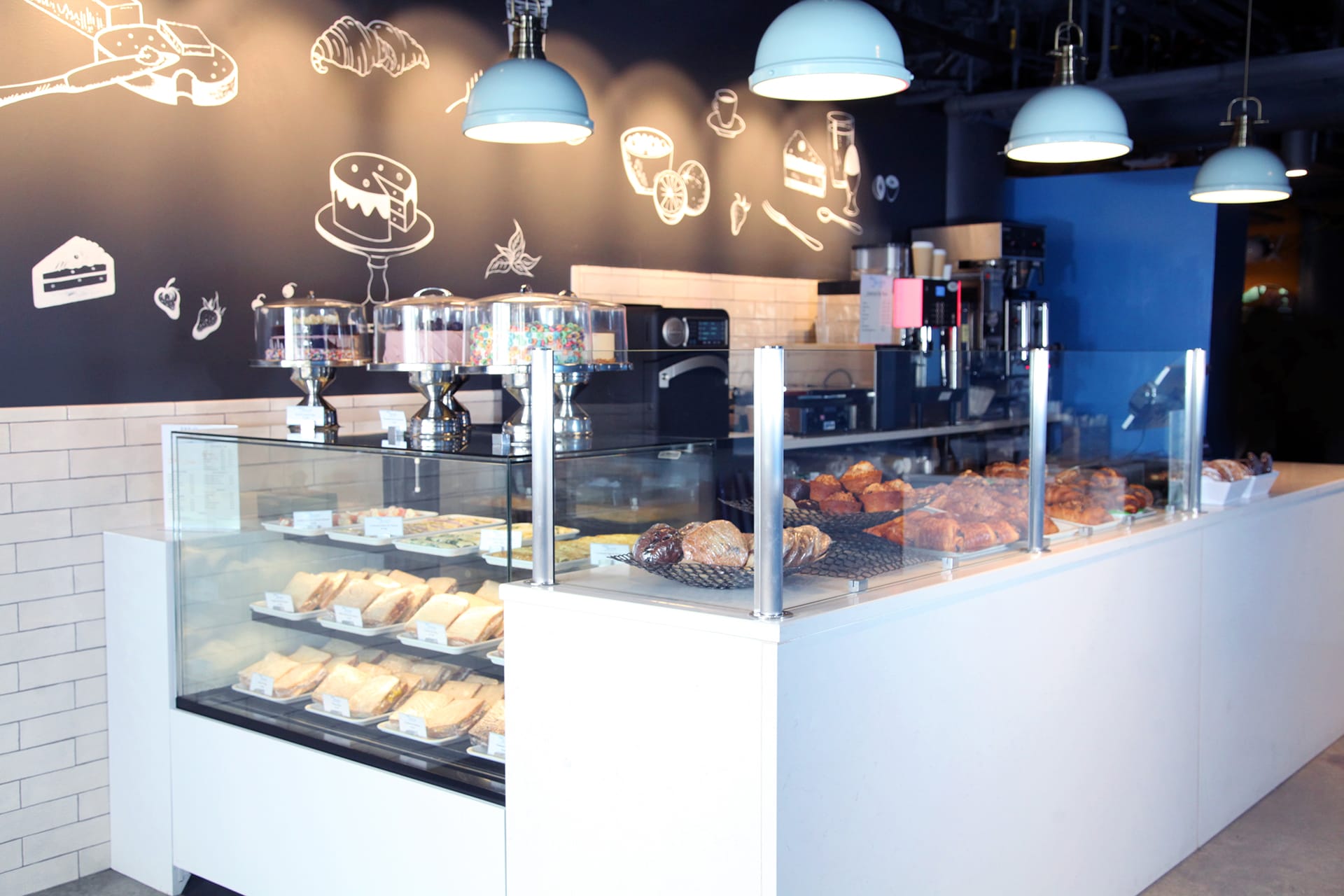 Counter with baked goods in a glass case, four powder blue pendant lights hanging above, and baked goods drawn in white on the black wall.