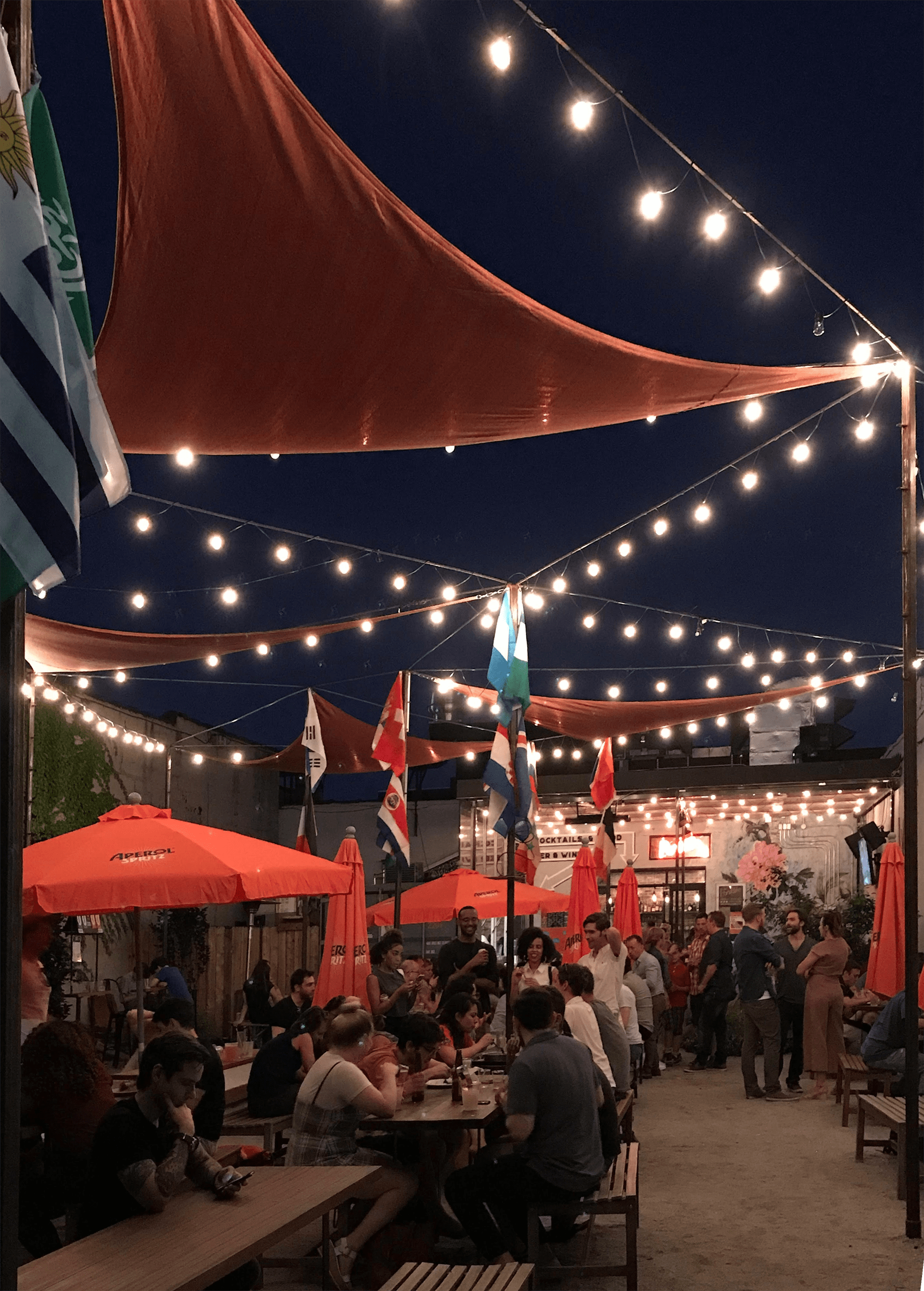 Courtyard of Parklife at night, with strings of lights, triangular orange awnings, and orange Aperol umbrellas. Groups of people sit at picnic tables and stand, talking and drinking.