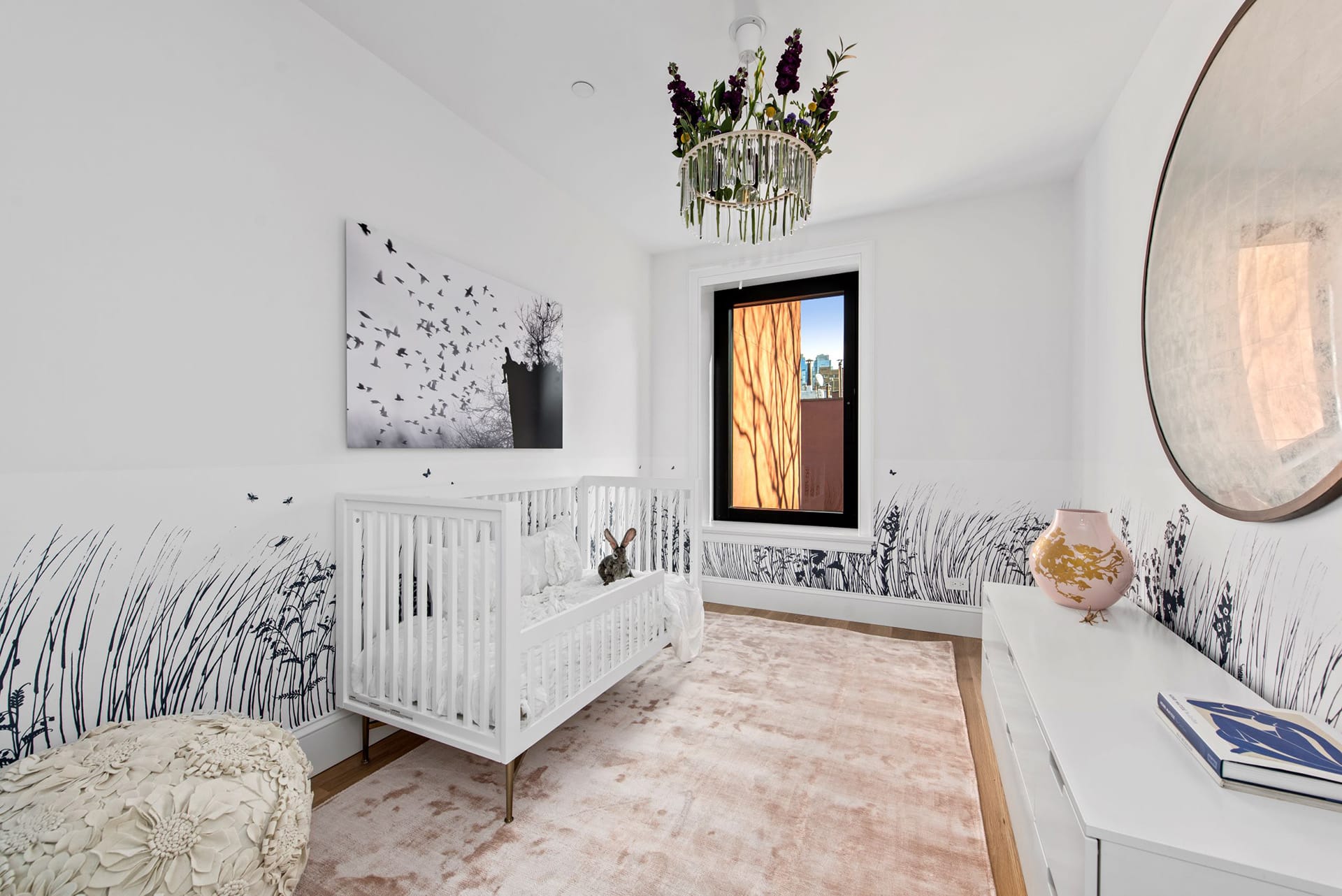 Nursery with a dusty rose carpet, white crib, wallpaper with wheat fields at the bottom, white dresser, circular mirror, flower-covered chandelier, and wood floors.