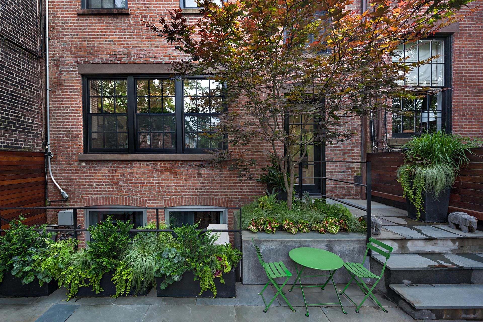 Rear façade of a brick house with a red tree, bluestone pavers, and green table and chairs